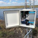 An open outdoor electrical box mounted on a metal pole by a body of water, containing various electronic components, wires, and a cylindrical device, with grass and blue sky in the background.