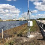 A weather monitoring station with sensors and antenna is installed next to a canal beside a road. The sky is partly cloudy, and there is grass and water nearby, with a building and more equipment in the background.