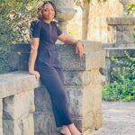 A woman in a black jumpsuit and heels leans against a stone wall in a sunlit garden with greenery and decorative stone urns in the background.