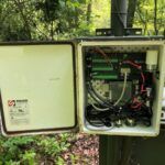 An open outdoor electrical control box with wiring and circuit boards inside, mounted on a metal post in a wooded area with green foliage surrounding it.