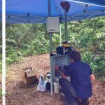 A person crouches under a blue canopy in a forested area, working with equipment in a metal box. Various bags, boxes, and tools are scattered on the ground nearby.