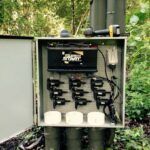 An open outdoor electrical control box with wiring, black battery labeled “Super Start,” multiple electrical components, and PVC piping, surrounded by dense green vegetation. Tools rest on top of the box.
