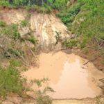 A muddy landslide area with a pool of brown water at the bottom, surrounded by broken trees and steep, eroded earth, with green vegetation at the top and edges.