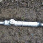 A water meter attached to a white PVC pipe partially buried in muddy soil with surrounding dirt and leaves visible.