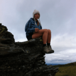 A person wearing a blue jacket, shorts, hiking boots, and a knit hat sits on the edge of a rocky outcrop, with cloudy skies and green landscape in the background.