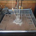 A scientific experiment setup with metal sensors and wires inserted into a cylindrical container surrounded by sand and cinder blocks, enclosed in a metal-framed box.