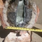 A partially melted glass bottle embedded inside a split rock, with a measuring tape stretched across to show its width, outdoors on a sunny day.