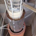 A scientific or industrial instrument with wires and metal components is placed inside a terracotta cylinder, surrounded by sandy material. Several cables extend from the top section, which is wrapped in insulation.