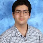 A young man with short dark hair and glasses, wearing a white and blue plaid shirt, poses in front of a blue textured backdrop.