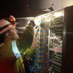 A worker in a hard hat and safety gear uses a flashlight to inspect or repair electrical wiring inside an open control panel in a dimly lit environment.