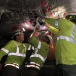 Three workers wearing safety gear and green reflective clothing install or repair equipment on a mesh-covered underground ceiling, illuminated by headlamps and surrounded by dark, rocky walls.