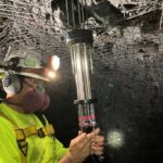 A worker in safety gear, including a helmet, headlamp, respirator, and ear protection, installs or inspects equipment with wires in an underground tunnel reinforced with metal mesh.