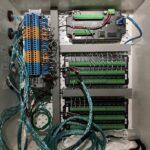 An open electrical control panel box contains neatly arranged terminal blocks, green circuit boards, black modules, and several bundles of blue wires connected to various components.