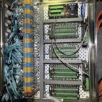 Metal electrical control panel open, showing organized blue and yellow crimped wires connected to terminal blocks and green circuit boards. Orange cables run outside the box on a dark floor.