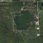 A satellite view of a forested area with a central lake, surrounded by numbered red markers labeled with codes, and intersected by dirt roads and clearings. Some buildings and fields are visible at the top right.