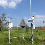A weather monitoring station with solar panels, sensors, and a pyramid-shaped device stands in a grassy field under a partly cloudy sky. Trees and a body of water are visible in the background.