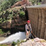 A person wearing climbing gear and a red helmet stands on a rocky ledge above a dam, secured by ropes, with water gushing from the dam and greenery visible in the background.