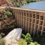 A large concrete dam releases water through its spillway into a rocky gorge below, surrounded by cliffs and greenery, with a calm reservoir behind the dam.