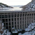 A large concrete dam is built between rocky, snow-covered hills, holding back a partially frozen reservoir. The dam features multiple vertical supports, and the landscape behind it stretches to the distant horizon.