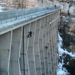 Two people are rappelling down the vertical face of a large concrete dam with snow-covered rocks below and on the sides. The dam is tall, with a fence along the top edge and a few people standing above.