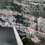 A concrete bridge spans a river, connecting rocky cliffs with green vegetation. The landscape features layered rock formations and slopes rising above the water.