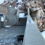 A concrete walkway with a railing leads to rocky stairs surrounded by snow. A solar panel is mounted on the left, and a black toolbox sits on the walkway. Rocky cliffs and patches of snow are in the background.