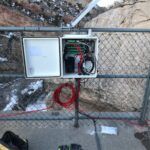 An open electrical or communications box with red wires and equipment is mounted on a chain-link fence outdoors near rocky cliffs. Tools, gloves, and a bag are on the concrete ground below. Snow patches are visible on the rocks.