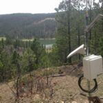 A weather monitoring station with solar panels stands on a grassy hillside overlooking a forested area and a lake surrounded by trees.
