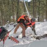 Two people wearing orange safety vests work together in a snowy forest, using a metal pole near an auger and a vehicle, surrounded by trees.
