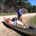 A man in a sun hat stands next to a canoe on a dirt road, holding a paddle. The canoe is filled with gear, backpacks, and supplies. Trees and greenery are visible in the background.