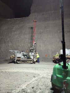 A worker in safety gear operates a drilling machine on tracked wheels inside a large, dimly lit underground cavern with high, rough walls. A green piece of equipment is visible in the foreground on the right.