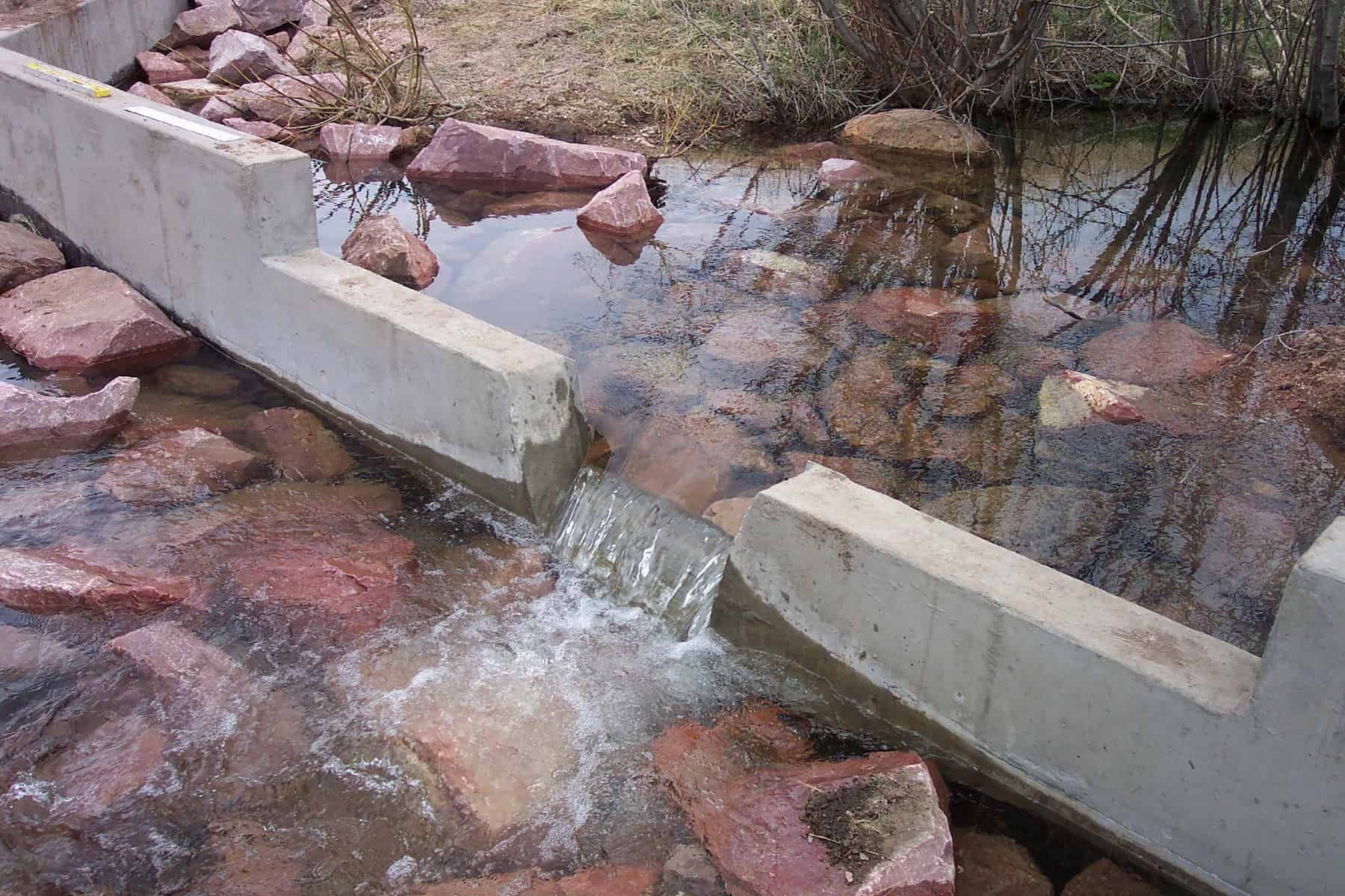 A small stream, part of mine water management efforts, flows between two concrete barriers, creating a mild waterfall over reddish rocks with some vegetation and dry grass visible on the banks.