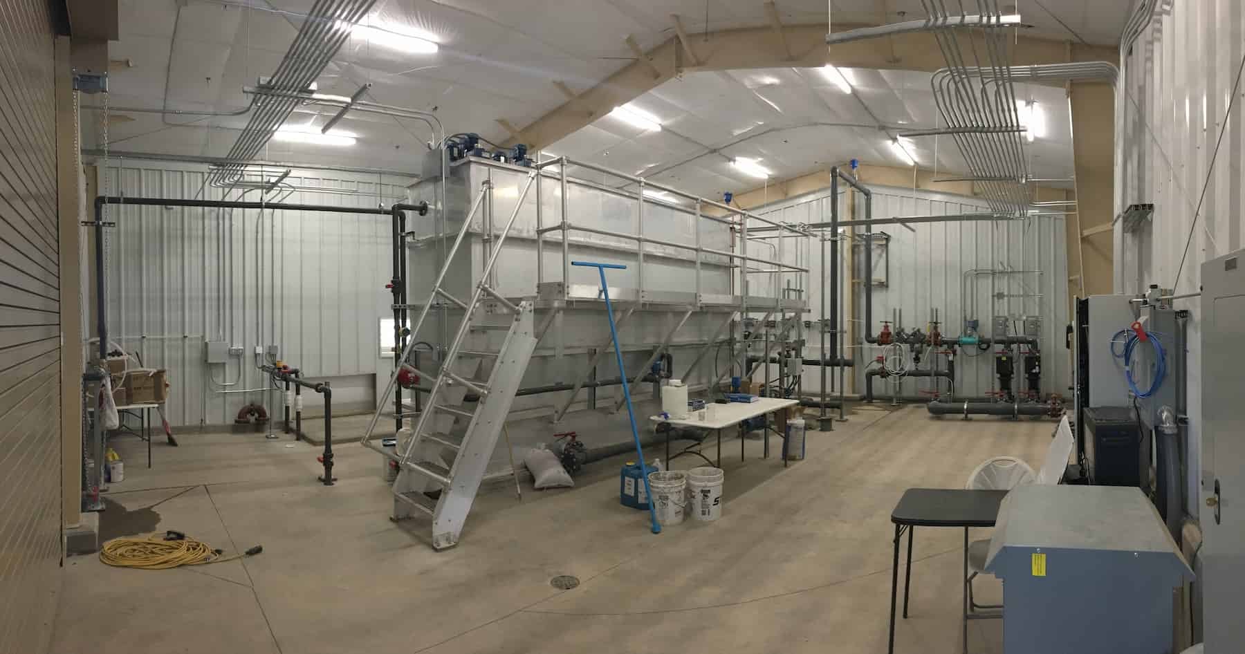 Large industrial room with high ceilings, featuring a metal platform with stairs, pipes, valves, and utility equipment essential for mine water management. Buckets, a broom, and tables with miscellaneous items are visible on the concrete floor.