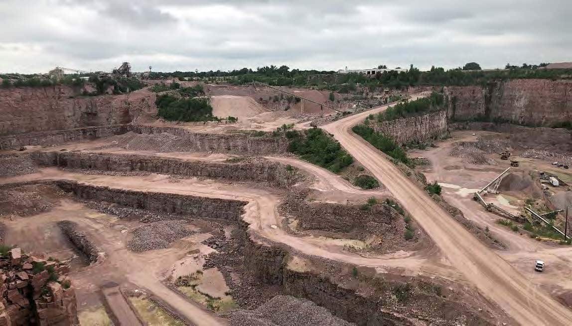 A large open-pit quarry with exposed rock layers, winding dirt roads, and sparse vegetation. Machinery and equipment are scattered throughout the site under a cloudy sky.