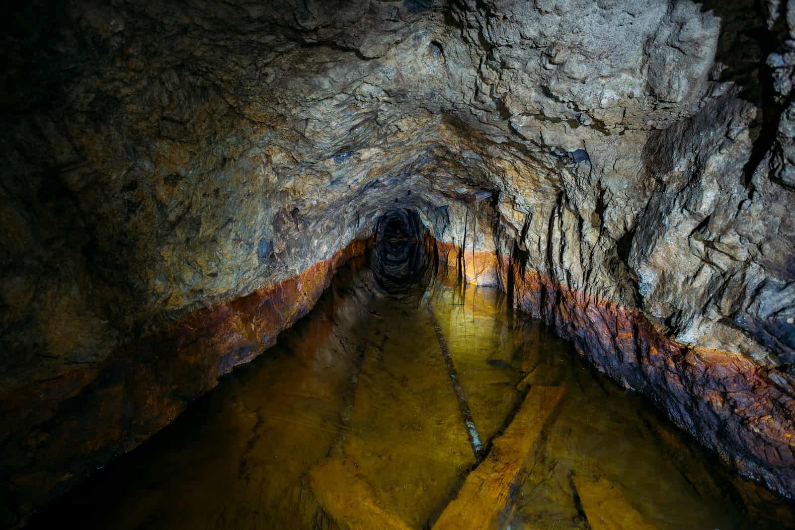 A dimly lit, rocky tunnel with jagged walls and a shallow, murky pool of yellowish water hints at the need for careful mine water management. Mineral streaks in orange and brown hues line the walls as the passage leads into darkness.