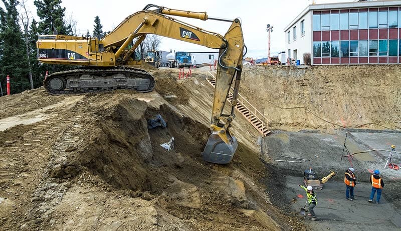A large yellow excavator digs into a steep dirt slope at a construction site, while several workers wearing safety gear stand below near a building and wooden stairs.