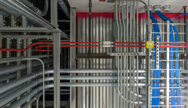 Rows of organized metal electrical conduits, red piping, and blue network cables run along an industrial wall and ceiling, showing a neatly arranged infrastructure system.