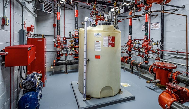 Large white chemical tank in a room with gray walls, surrounded by red pipes, valves, gauges, and control panels. The area appears clean and organized, likely part of an industrial or fire protection system.