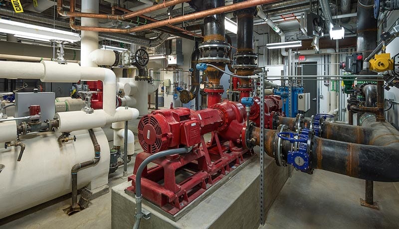 A mechanical room with large red pumps, white insulated pipes, black and blue valves, and various metal pipes and fittings, all set on a concrete floor with overhead lighting.