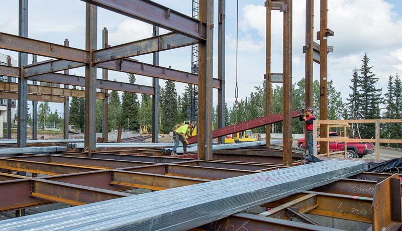 Two construction workers are installing steel beams on a building framework at a construction site, surrounded by trees and construction vehicles under a partly cloudy sky.