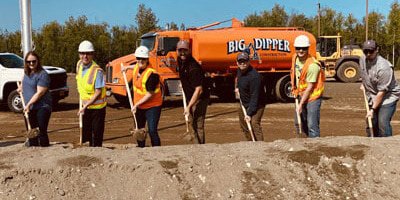 Seven people wearing safety gear hold shovels at a construction site during a groundbreaking ceremony, with an orange Big Dipper truck and construction equipment in the background.