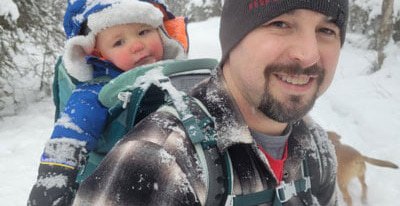 A man wearing a beanie carries a bundled-up baby in a hiking backpack through snowy woods. Both have snow on them, and a dog is partially visible in the background.