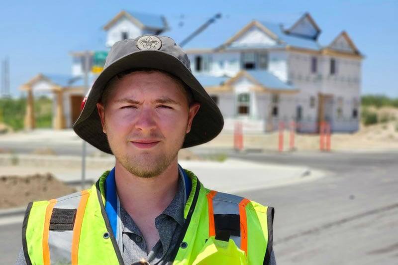 A person wearing a wide-brim hat and safety vest stands outside in front of a partially constructed house on a sunny day.