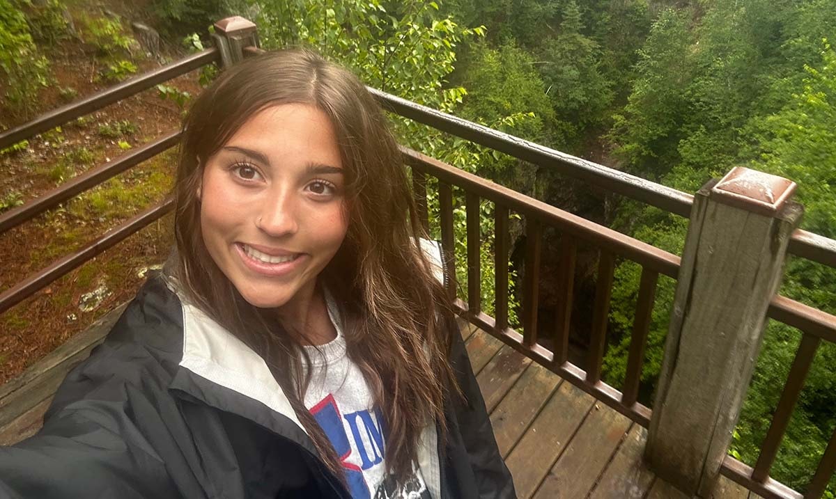 A young woman in a black rain jacket smiles for a selfie on a wooden deck surrounded by lush green trees in a forested area.