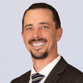 A smiling man with short brown hair and a goatee, wearing a dark suit, white shirt, and striped tie, poses against a light gray background.