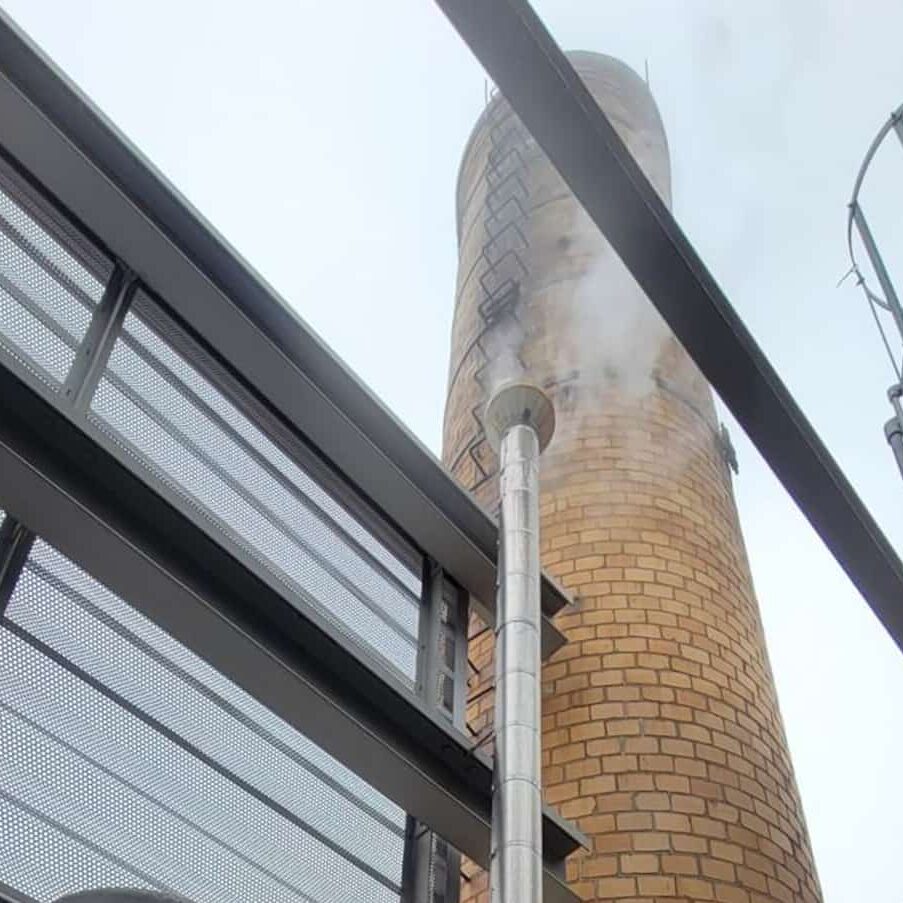 A view looking up at a tall brick chimney emitting white smoke, surrounded by metal structures and safety railings against a cloudy sky.