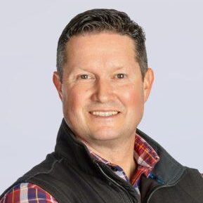 A man with short dark hair smiles at the camera. He is wearing a plaid shirt under a dark vest, and the background is light gray.