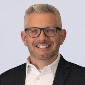 A smiling man with short gray hair and a trimmed beard wearing black glasses, a white collared shirt, and a dark suit jacket, against a plain light background.