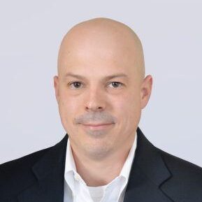 A man with a bald head, light skin, and brown eyes, wearing a white collared shirt and a dark blazer, is facing forward and smiling slightly in front of a plain light gray background.