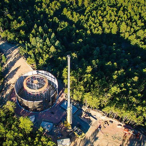 Aerial view of a construction site with a large circular structure surrounded by dense forest and construction equipment, including a tall crane, visible on a cleared area next to the trees.